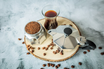 Top-angle rustic setup: moka-pot lid, basket of ground coffee and cup of black coffee on a wood slice, roasted beans scattered across pale concrete background