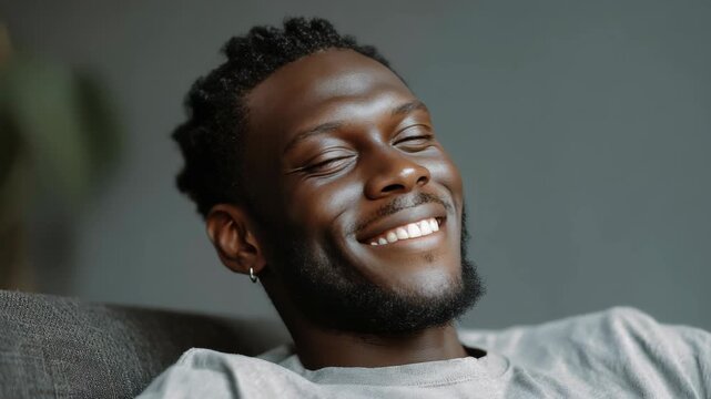 A cheerful young man with a beard and dreadlocks, wearing a gray t-shirt, smiling broadly with his eyes closed in an expressive gesture of joy.