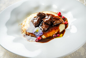 Gourmet meat dish in rich sauce with mashed potatoes, red berries, and edible flower, elegantly plated on white dish over decorative tablecloth, perfect for upscale dining themes.