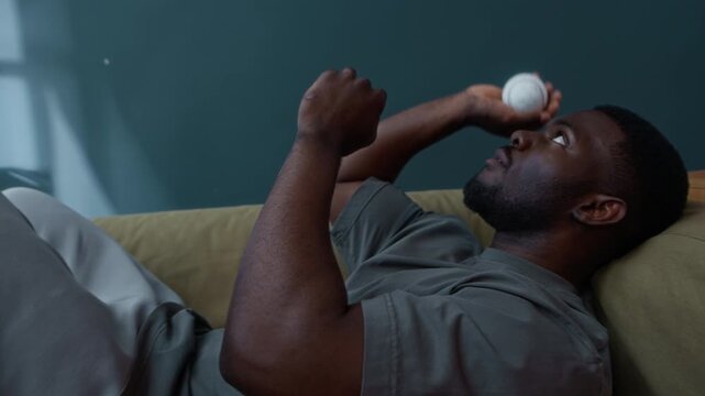 Bored young African American man under house arrest wearing electronic tag lying on couch and throwing ball trying to kill time on probation