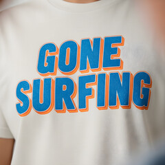 Close-up of a white t-shirt featuring a bold, three-dimensional "Gone Surfing" graphic in blue and orange.