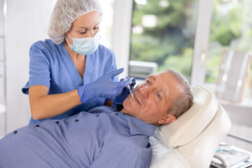 Experienced female cosmetologist working with senior male client in clinic of aesthetic medicine, performing minimally invasive procedure using filler injections to correct lips contour