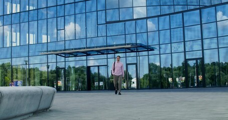 An elegant business man walks along a city street past an office building. The backpack is on his shoulder.