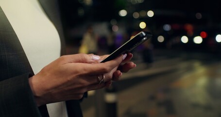 A woman uses a mobile phone while standing on a city street in the evening. Close-up of her hands, unrecognizable person