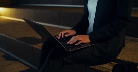 A woman uses a laptop while sitting on the steps of a city street at night. Close-up of her hands, unrecognizable person