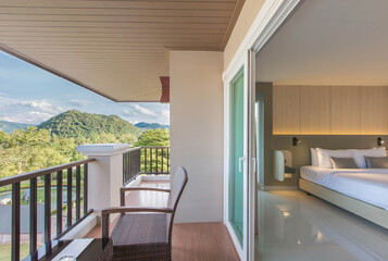 Balcony with Mountain View, A cozy balcony with wicker chairs and a glass table overlooking lush green forests and distant mountains under a cloudy blue sky.