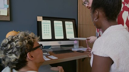 Focused diverse team of UX designers sitting at desk with computer displaying mobile app interface on screen discussing wireframe pointing at sketch in notebook while collaborating at office - Powered by Adobe