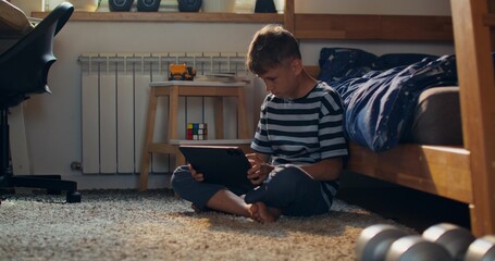 The boy is intently using a tablet while sitting on the floor next to the bed