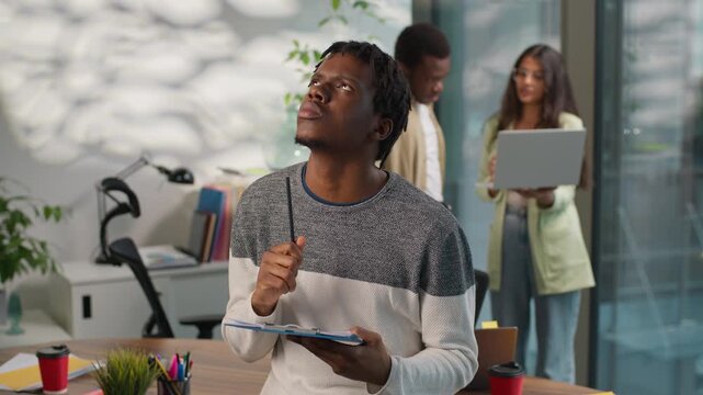 Pensive thinking African American informal business man businessman male guy startup owner writing notes in office on background of multiracial colleagues working with laptop talking multiracial team