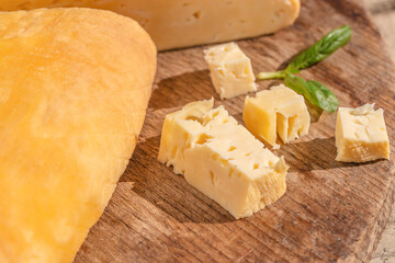 Small cubes of smoked cheese rest on a rustic wooden board beside a large cheese wedge and fresh green basil leaves in warm sunlight
