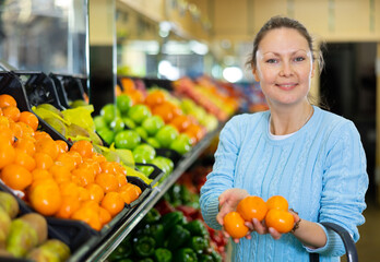 Glad middle-aged woman customer buying fresh mandarins at the counter in big greengrocery