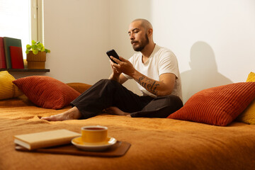 Man relaxing on a bed with a phone, next to a book and a cup of tea.
