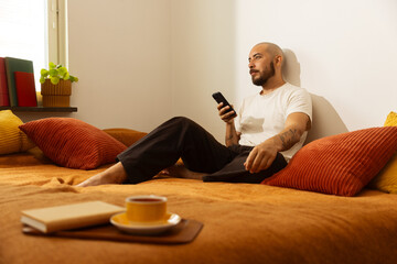 Man relaxing on a bed with a phone, next to a book and a cup of tea.
