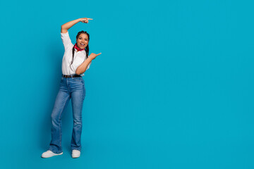 Portrait of a cheerful young woman in casual attire pointing playfully against a vibrant blue...