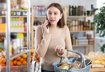 Positive young woman looking for something, holding cart with food products and talking on mobile...