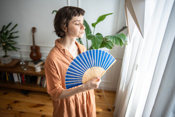 Frustrated exhausted woman fanning herself struggling with sweltering weather, feel heat dehydration. Airless apartment with one window, health deterioration, vulnerability to hot weather, no AC