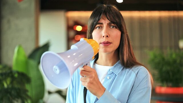 Passionate businesswoman, wearing a blue shirt, powerfully speaks into a megaphone indoors. Her serious expression conveys an important, urgent announcement.
