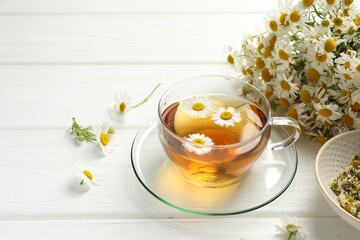 Aromatic tea in glass cup and chamomile flowers on white wooden table, closeup