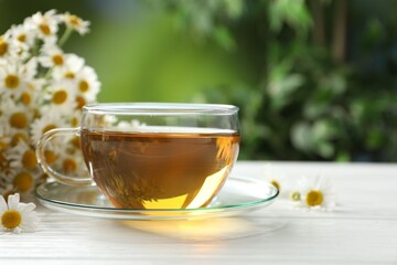 Aromatic tea in glass cup and chamomile flowers on white wooden table, closeup