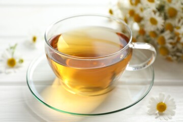 Aromatic tea in glass cup and chamomile flowers on white wooden table, closeup