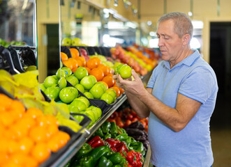 in modern supermarket, elderly man carefully selects apples and puts them in shopping cart