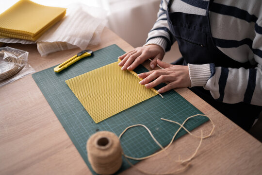 Hispanic craftswoman in apron making candles from natural wax sheet in workshop, close-up.