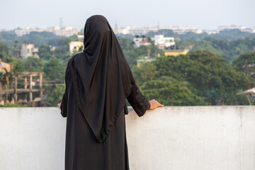 Solo Bengali Muslim woman in black hijab stands on a rooftop, back to viewer, contemplating the...