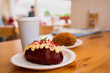 A delicious red velvet pastry topped with cream cheese frosting sits on a plate beside a takeaway coffee cup, ready to be enjoyed at a café.