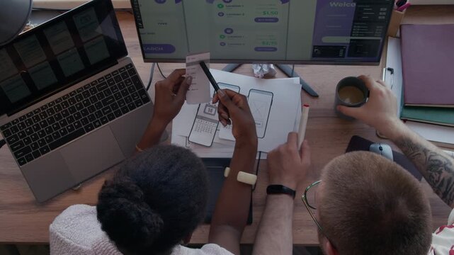 Top view of Black female UI designer pointing at mobile app wireframe on paper talking to male IT programmer developing user interface collaboratively at office desk with computer