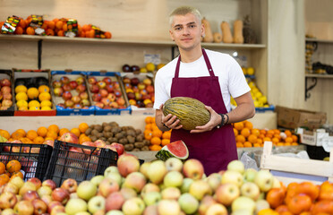 Guy seller with watermelon in hand stands in trading hall of vegetable store, wearied in apron stands in trading hall of minimarket, ready to greet visitors and offer food stuff. .