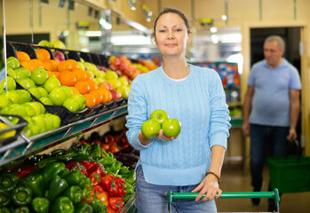 Fototapeta premium Pleased middle-aged woman buyer purchasing fresh apples at the counter in large vegetable shop