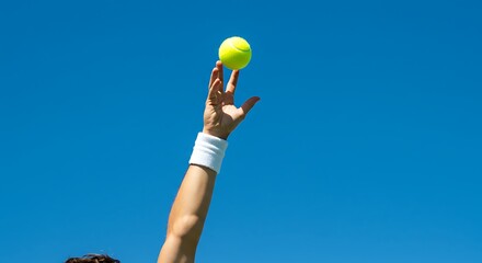 A tennis player's hand tosses a yellow ball into the air, preparing to serve against a clear blue sky.