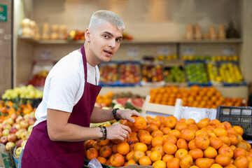 While working in shop, employee forms display case, puts ripe imported mandarin in pile. Open display of goods in self-service shop. Male employee makes attractive display case with vegetables..