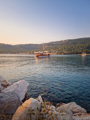 Tranquil harbor scene at dawn and a wooden boat with mast anchored in the calm sea water. Green hills rise across the bay bathed in the warm morning light. Moonlight beach in Kemer, Turkey