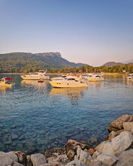 Morning light bathes the Moonlight beach in Kemer, Turkey. Boats rest in the calm bay with turquoise Mediterranean sea water. Lush green hills with palm trees seen at horizon
