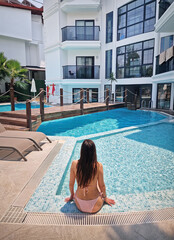 Beautiful woman in a pink bikini sits relaxed by a resort pool, gently splashing the water. The serene setting features a white hotel building, and yard with palm trees, and a wooden bridge