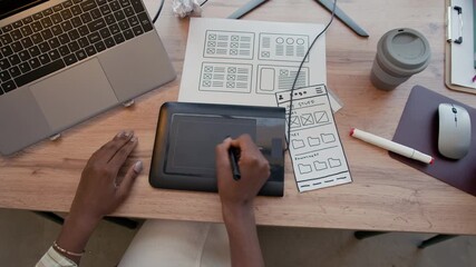 Top view of African American woman drawing on graphics tablet with digital pen designing UI of mobile app while working on desktop cluttered with paper sketches at office - Powered by Adobe