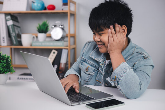 Young Asian boy smiling while using a laptop at his desk, representing successful online learning, engaging educational content, and positive digital experiences for children.