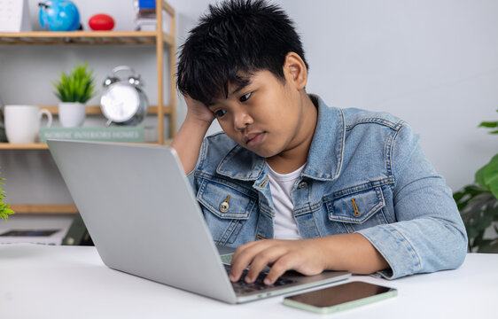 Young Asian boy in denim jacket looking bored and exhausted while using laptop, representing online learning fatigue, screen time issues, and study stress in children. - Powered by Adobe