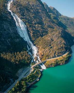 Aerial view of the cascading Langfoss waterfall plunging down a rugged mountain into the turquoise fjord, contrasted by the road below, Fj&Atilde;&brvbar;ra, Vestland, Norway.