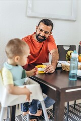 Morning moment with father feeding baby boy in high chair with a spoon. Smiling dad interacts with...
