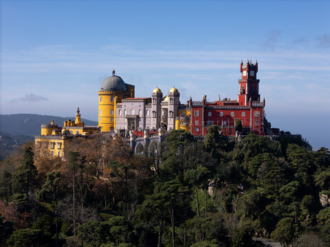 Sintra, Portugal - 11 June 2024: Aerial view of Pena Palace atop a verdant hill, its vibrant yellow, red, and grey walls contrasting with the deep green forest below.