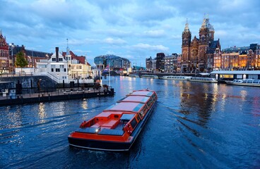 A tour boat cruising on the canal near Amsterdam's Central Railway Station, with the spires of Saint Nicholas Basilica towering in background and city lights reflected in the water at blue dusk