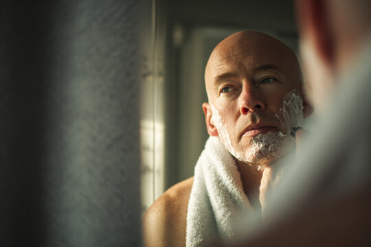 Man shaves his face, looking in mirror. Depicts grooming routine. Useful for skincare ads, male hygiene concepts, and self-care marketing.