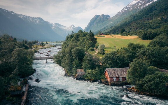 Aerial view of the vibrant turquoise river rushing beneath the bridge, juxtaposed with the lush green forests and snow-capped mountains, Olden, Vestland, Norway.