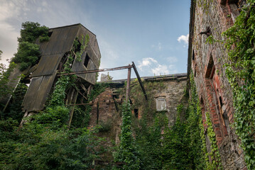 Ruins of an abandoned, industrial building shows crumbling stone and brick, with boarded-up windows and ivy climbing the surface. Photo taken in Inniskeen, County Monaghan, Ireland