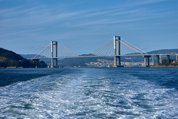 Sea wake in the foreground as a boat moves away, leaving Rande Bridge behind in the Vigo Estuary on a clear day