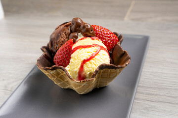 Ice cream dessert with vanilla, chocolate, strawberries, syrup, and chocolate chips in a waffle bowl on a gray rectangular plate over a wooden surface.
