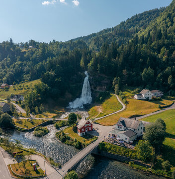Aerial view of Steinsdalsfossen waterfall cascading down lush green slopes, contrasting with the quaint village below and the clear river, Norheimsund, Vestland, Norway.