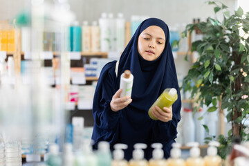 Muslim woman carefully chooses a natural shampoo and conditioner for her hair at a pharmacy....